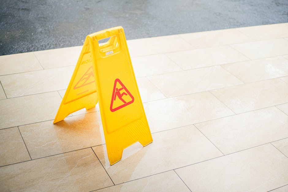 A yellow wet floor warning sign with a red triangular symbol indicating a slip hazard, placed on a light beige tiled floor near the entrance of a property. The tiles are clean and evenly laid, with subtle grout lines visible. In the background, a section of dark asphalt driveway or street can be seen, suggesting the location is just outside the property. The image is well-lit with natural light, capturing the area where professional removals, such as packing and home relocation efforts by Man and Van Redbridge, might take place. The sign is positioned to alert movers or residents to exercise caution while walking or carrying furniture during the loading process involved in house removals or furniture transport.