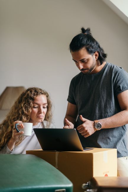 A man with long dark hair tied in a bun and wearing a grey t-shirt is standing indoors next to a woman with curly blonde hair who is sitting at a table. The woman holds a white mug in her right hand and looks at an open laptop placed on a cardboard box in front of her. The setting appears to be inside a house, with a light-colored wall and a lamp in the background. They are engaged in a discussion related to packing or planning a home relocation, which is relevant to moving and removal services. The environment suggests a residential space used for planning or organizing a move, highlighting the process of packing, furniture handling, or logistical coordination during house removals.