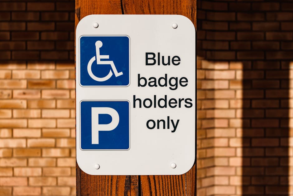 A rectangular white parking and accessibility sign mounted on a wooden post in front of a brick building. The top section of the sign displays a blue wheelchair symbol indicating disabled access, alongside the text 'Blue badge holders only' in black letters. The lower section features a large blue letter 'P' symbol, indicating parking availability. The overall scene appears to be outside a residential or commercial property, with the sign positioned on a pavement or driveway area, and natural daylight illuminating the setting. This image relates to parking regulations that can affect home relocation and furniture transport logistics, as managed by companies like Man and Van Redbridge during house removals and moving processes.