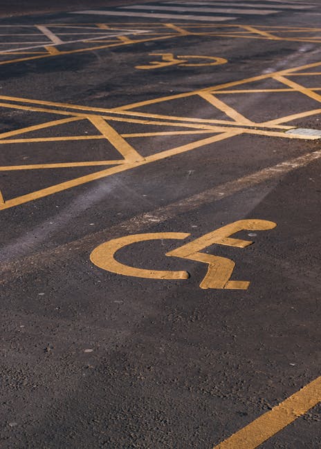 A close-up view of a parking lot surface showing two yellow wheelchair symbols painted on the dark asphalt, indicating designated accessible parking spaces. The symbols are positioned next to each other, with one in the foreground and the other further back. The lines marking the parking bays are slightly worn but clearly visible, forming a grid pattern with diagonal lines and vertical boundaries. The surface appears clean with minimal debris and some slight texture from the asphalt material. The image captures the clarity of the parking space markings intended for individuals with mobility impairments, relevant to house removals and parking permit advice as offered by Man and Van Redbridge, which specializes in house removals and moving logistics within the Redbridge area.