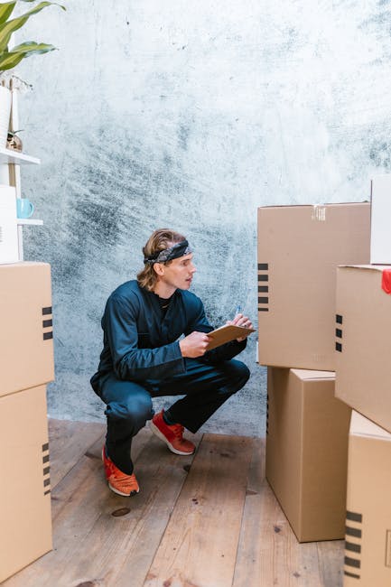 A man wearing a black headband, navy blue jumpsuit, and orange sneakers is crouching down inside a room with a textured light blue wall and wooden flooring. He is holding a clipboard and appears to be
