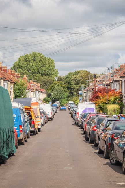 A residential street during daytime with numerous parked cars lining both sides of the road, including a mix of sedans, hatchbacks, and vans, some of which belong to various local service providers. T