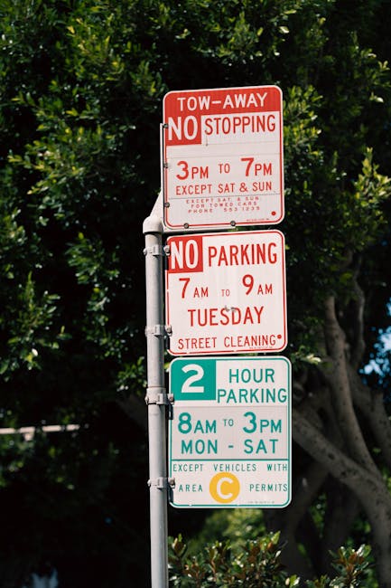A residential street during daytime with numerous parked cars lining both sides of the road, including a mix of sedans, hatchbacks, and vans, some of which belong to various local service providers. T