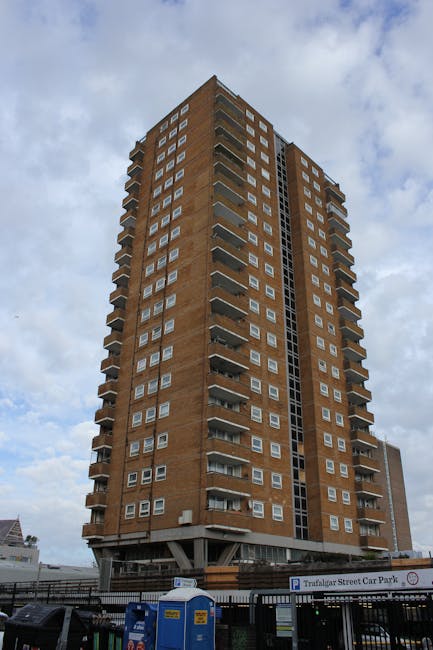 A tall multi-storey residential building constructed from brown brick, with numerous small rectangular windows arranged in a grid pattern. Several small balconies with metal railings are visible on the front and sides of the building, some with furniture or decoration. The building is situated on a paved street with a parking area in front, including a sign indicating 'Trafalgar Street Car Park' and a row of blue portable toilets. The low-level base of the building appears to include commercial or shared facilities. Natural daylight illuminates the scene, with some clouds in the sky, and the overall setting suggests an urban environment suitable for residential home relocations or furniture transport services, as provided by Man and Van Redbridge.