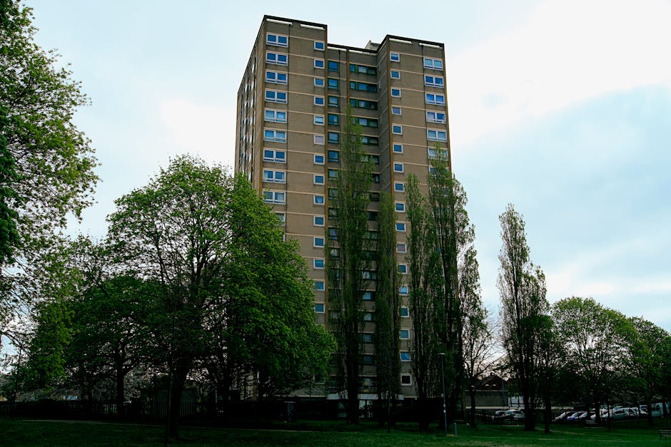 A tall, multi-storey residential building with numerous windows, situated in an urban environment surrounded by several mature trees with green foliage. The building appears to be constructed of concrete with a modern high-rise design. The sky overhead is partly cloudy, providing diffuse natural light. In the foreground, a grassy area with pathways and parked cars can be seen, indicating a residential setting with accessible communal outdoor space. This image is associated with house removals and relocation services, as it portrays a typical apartment block that may be involved in home relocation or furniture transport processes. Man and Van Redbridge might utilize such images on their website to illustrate large-scale residential moves or complex packing and loading operations for flats in this type of building.