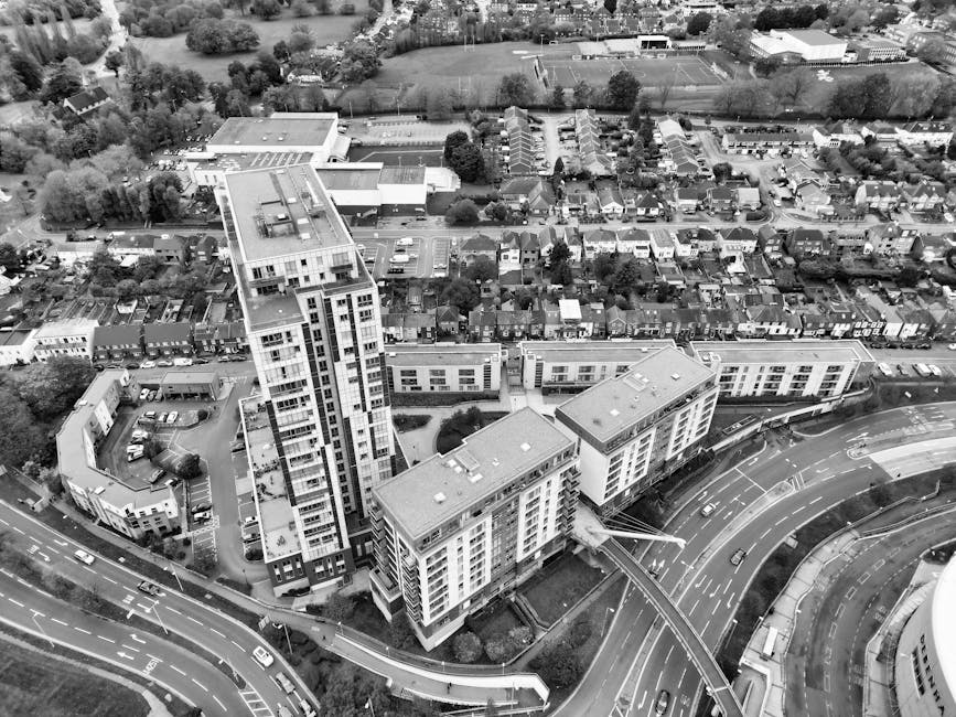 A tall multi-storey residential building constructed from brown brick, with numerous small rectangular windows arranged in a grid pattern. Several small balconies with metal railings are visible on the front and sides of the building, some with furniture or decoration. The building is situated on a paved street with a parking area in front, including a sign indicating 'Trafalgar Street Car Park' and a row of blue portable toilets. The low-level base of the building appears to include commercial or shared facilities. Natural daylight illuminates the scene, with some clouds in the sky, and the overall setting suggests an urban environment suitable for residential home relocations or furniture transport services, as provided by Man and Van Redbridge.