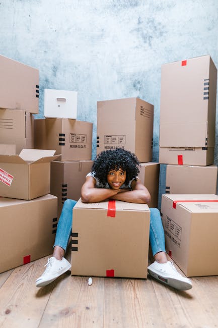 A young woman with curly black hair and a bright smile is sitting on the floor inside a room filled with numerous cardboard moving boxes of various sizes, some partially open with packing paper visible inside. The boxes are sealed with red and clear packing tape, and are stacked in several directions around her. She is wearing a white top, blue jeans, and white sneakers. The room has a light blue, textured wall and a wooden floor, indicating a home in the process of relocation. The woman appears relaxed, possibly taking a break during the packing and moving process, which is part of a house removal or furniture transport operation. The image conveys a busy, organized moving environment, typical of professional removals including services offered by Man and Van Redbridge, supporting efficient packing and home relocation.