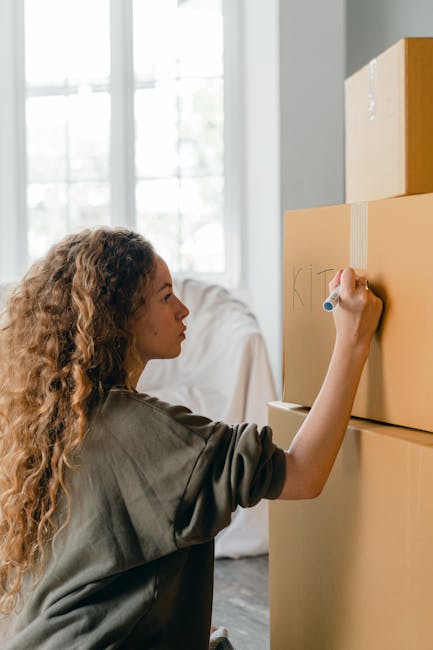 A young woman with curly black hair and a bright smile is sitting on the floor inside a room filled with numerous cardboard moving boxes of various sizes, some partially open with packing paper visible inside. The boxes are sealed with red and clear packing tape, and are stacked in several directions around her. She is wearing a white top, blue jeans, and white sneakers. The room has a light blue, textured wall and a wooden floor, indicating a home in the process of relocation. The woman appears relaxed, possibly taking a break during the packing and moving process, which is part of a house removal or furniture transport operation. The image conveys a busy, organized moving environment, typical of professional removals including services offered by Man and Van Redbridge, supporting efficient packing and home relocation.
