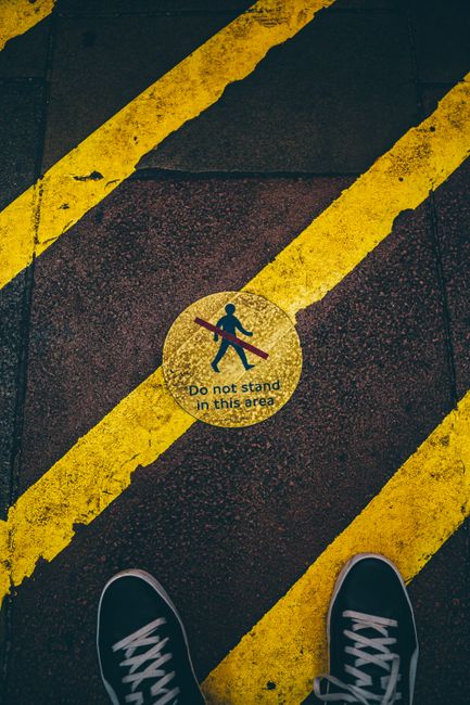 The image shows a close-up view of a paved ground surface marked with yellow and black diagonal stripes, typically indicating a designated loading or restricted area. In the center, there is a circular yellow sign with black text and a symbol: a person walking with a red slash crossing over the figure, accompanied by the message 'Do not stand in this area', indicating a safety or no-standing zone. At the bottom of the image, the toes of a person's shoes are visible, positioned near the sign, suggesting the individual is standing just outside or on the edge of the marked area. The surface appears to be outdoors, with lighting indicating daytime. This setting reflects a controlled space relevant to home relocation or furniture transport, consistent with the services provided by Man and Van Redbridge as seen on their website, and is useful in illustrating guidelines for safe loading and unloading during house removals.