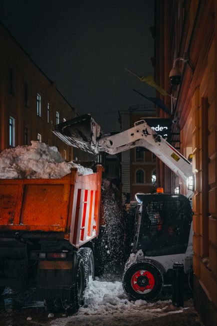 A compact skid-steer loader with a white arm and black body, equipped with a large bucket attachment, is in the process of loading snow and ice from a small orange tipper truck parked on a narrow street at night. The tipper truck is filled with snow, some of which is spilling over the sides as the snow is being lifted out. The scene is illuminated by the loader’s work lights, casting light on the snowy ground and surrounding buildings. To the right, a wooden building with vertical paneling is visible, with a small overhang and some mounted equipment. In the background, multi-storey residential or commercial buildings can be seen, with windows and signage. The environment suggests a winter cleaning or snow removal operation, relevant to house removals and logistics services provided by Man and Van Redbridge, situated in the context of home relocation and furniture transport, with a focus on efficient handling of winter weather conditions.