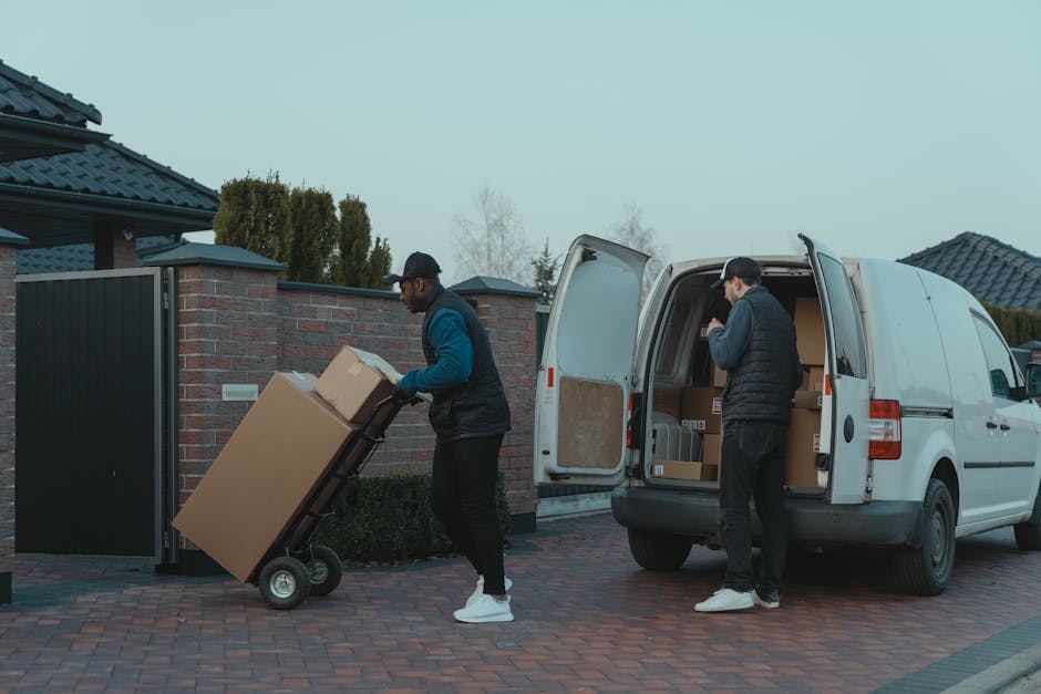 Two removal men from Man and Van Redbridge are engaged in a home relocation task outdoors on a paved driveway in South Woodford, E18. One man, dressed in a black cap, blue hoodie, and black trousers, is pushing a two-wheeled hand truck loaded with large cardboard boxes towards a white panel van. The other man, wearing a black cap, dark jacket, and dark trousers, is standing near the open rear doors of the van, organizing or preparing additional boxes for loading into the vehicle. The van's interior is visible and contains multiple packed cardboard boxes and other packaging materials, indicating an ongoing packing and moving process. In the background, residential houses with brick and tiled roofs are partially visible, along with trees and a brick wall, under an overcast sky, reflecting typical urban moving logistics. This scene captures the effective execution of packing, furniture transport, and loading activities associated with professional removals services found on [DOMAIN], aligned with the house and flat relocation services in the South Woodford area.