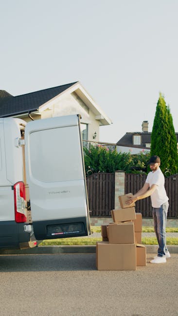 A man wearing a white t-shirt and jeans is standing beside a white panel van with its rear door open, on a residential street with a partly cloudy sky. He is stacking or unpacking three cardboard boxes of various sizes, some placed on the pavement and others being lifted or moved. The boxes appear to be used for packing belongings during a home relocation, with the van positioned close to the curb, ready for loading or unloading. In the background, there is a white house with a dark roof, a green hedge, and a tall coniferous tree, indicating a suburban environment. This scene illustrates the process of furniture transport and packing and moving logistics, typical of services offered by Man and Van Redbridge, especially related to house removals in South Woodford, E18, Redbridge.