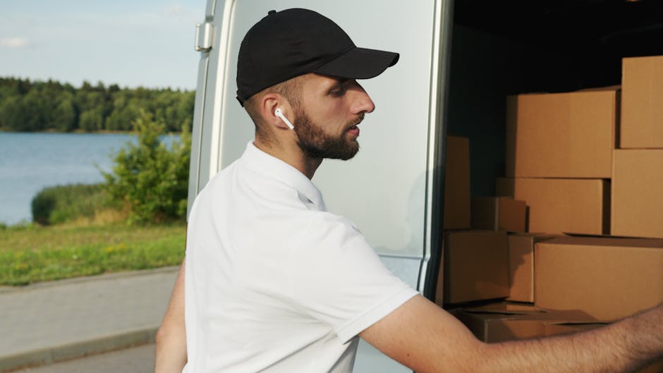 Two removal men from Man and Van Redbridge are engaged in a home relocation task outdoors on a paved driveway in South Woodford, E18. One man, dressed in a black cap, blue hoodie, and black trousers, is pushing a two-wheeled hand truck loaded with large cardboard boxes towards a white panel van. The other man, wearing a black cap, dark jacket, and dark trousers, is standing near the open rear doors of the van, organizing or preparing additional boxes for loading into the vehicle. The van's interior is visible and contains multiple packed cardboard boxes and other packaging materials, indicating an ongoing packing and moving process. In the background, residential houses with brick and tiled roofs are partially visible, along with trees and a brick wall, under an overcast sky, reflecting typical urban moving logistics. This scene captures the effective execution of packing, furniture transport, and loading activities associated with professional removals services found on [DOMAIN], aligned with the house and flat relocation services in the South Woodford area.